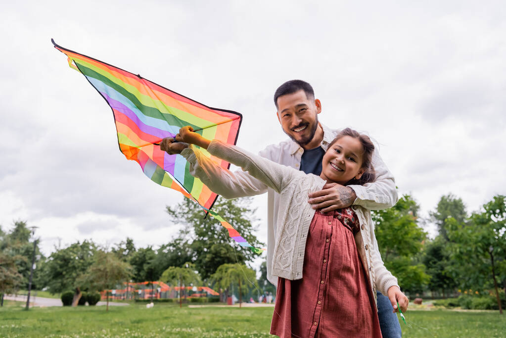 Tattooed asian dad hugging daughter with flying kite and looking at camera in park  - Foto, imagen