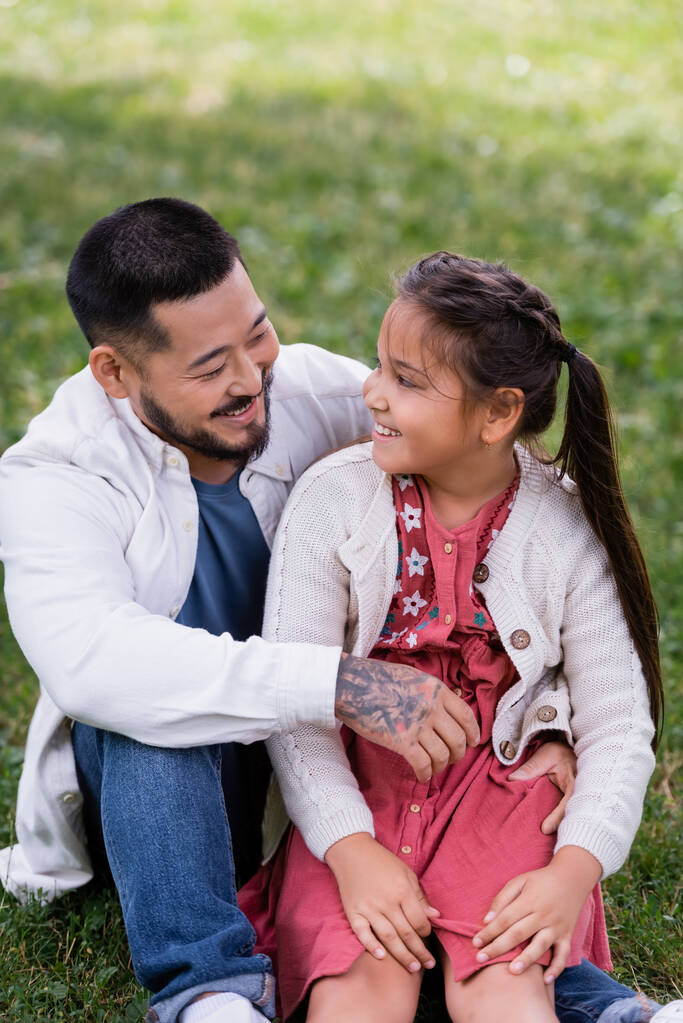 Τατουάζ Ασιάτης γονέας αγκαλιάζει την κόρη στο γρασίδι στο πάρκο  - Φωτογραφία, εικόνα