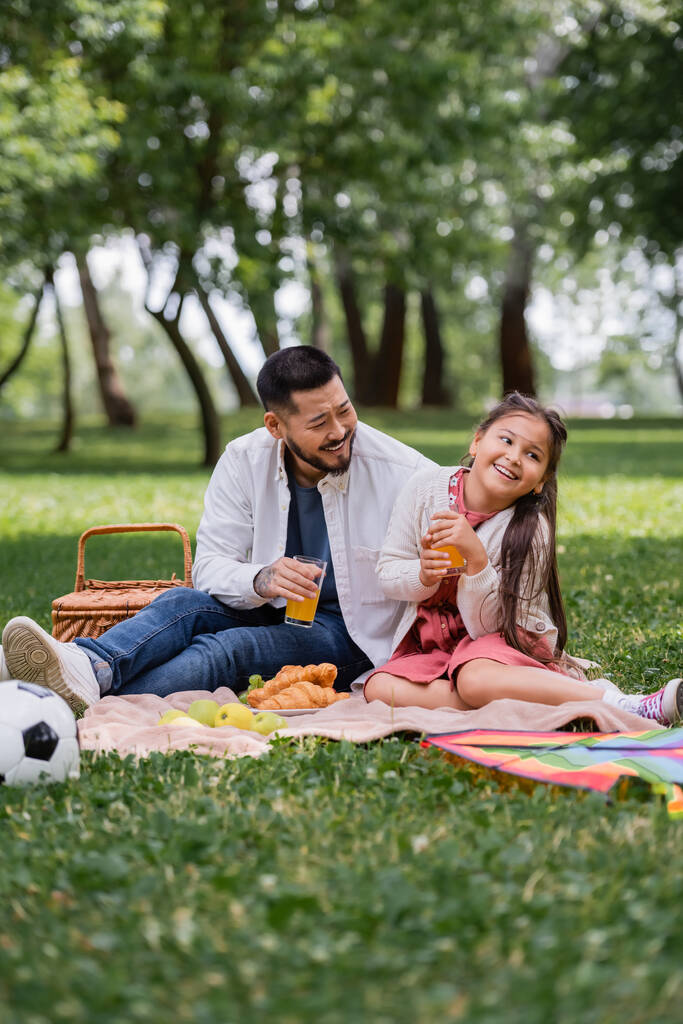 Счастливая азиатская семья держит апельсиновый сок рядом с едой и футболом в летнем парке  - Фото, изображение