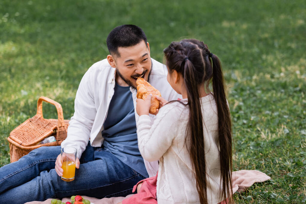 Child holding croissant near asian dad with orange juice in park  - Foto, afbeelding