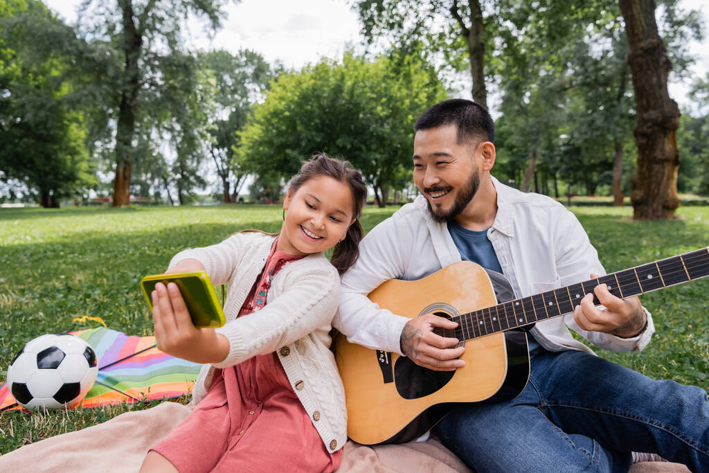 Cheerful asian girl taking selfie with dad playing acoustic guitar near flying kite and football in park  - Foto, afbeelding