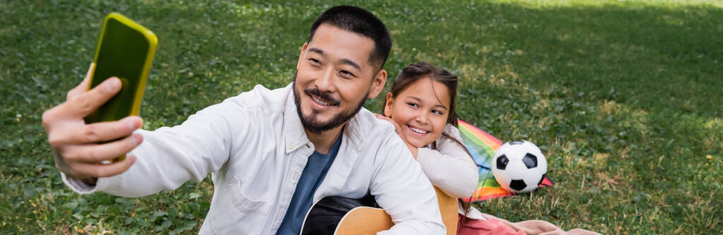 Asian dad taking selfie on cellphone while holding acoustic guitar near child in park, banner  - Photo, image