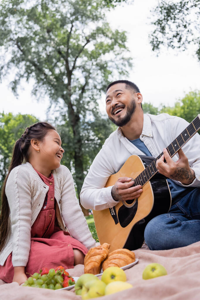 Positive asian parent playing acoustic guitar near daughter and blurred food in park  - Photo, Image