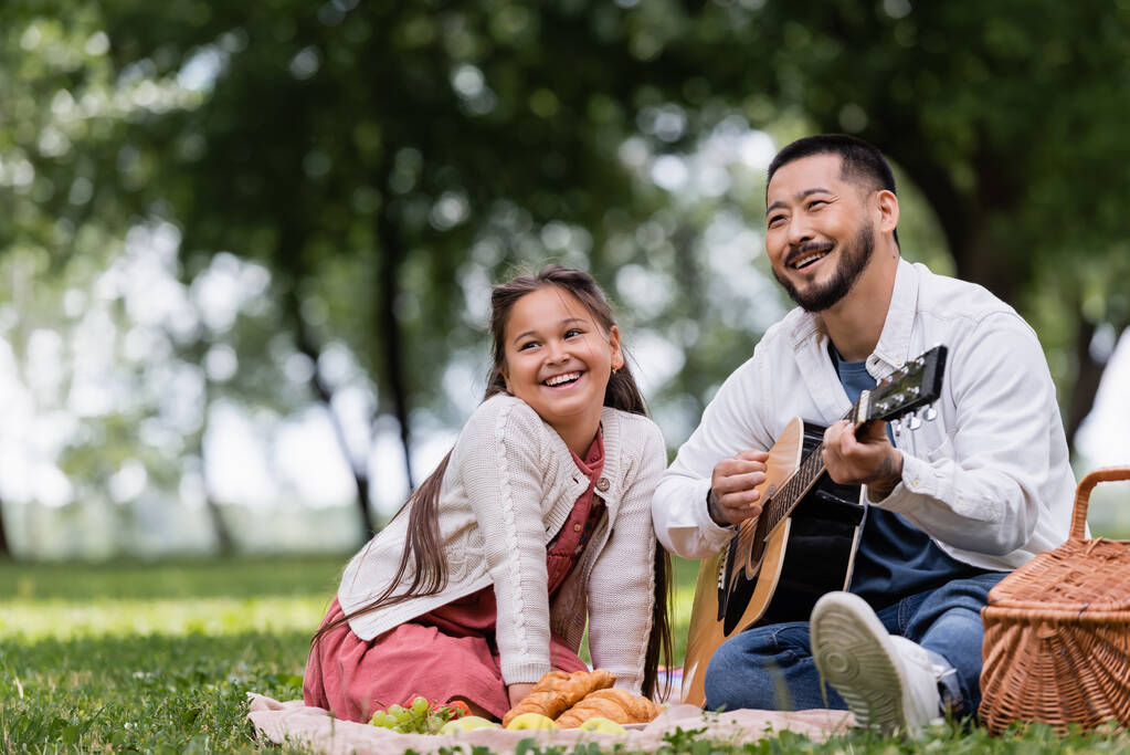 Улыбающийся азиатский папа играет на акустической гитаре рядом с дочерью и круассанами на одеяле в парке  - Фото, изображение