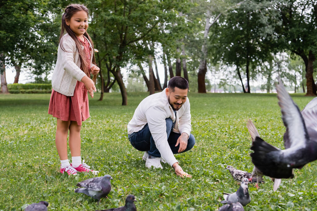 Позитивная азиатская семья кормит голубей на газоне в летнем парке  - Фото, изображение