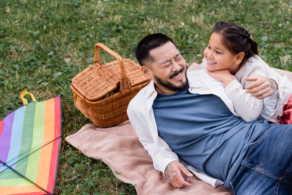 Happy asian dad lying near daughter and wicket basket in park  - Zdjęcie, obraz