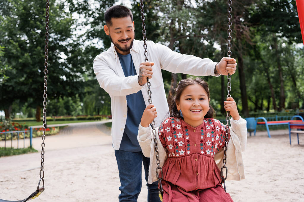 Smiling father standing near asian daughter on swing in park  - 写真・画像