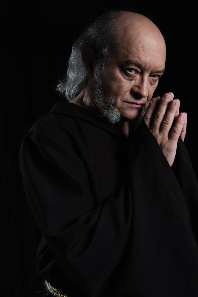 poważny mnich patrząc w aparat podczas modlitwy odizolowany na czarny - Zdjęcie, obraz