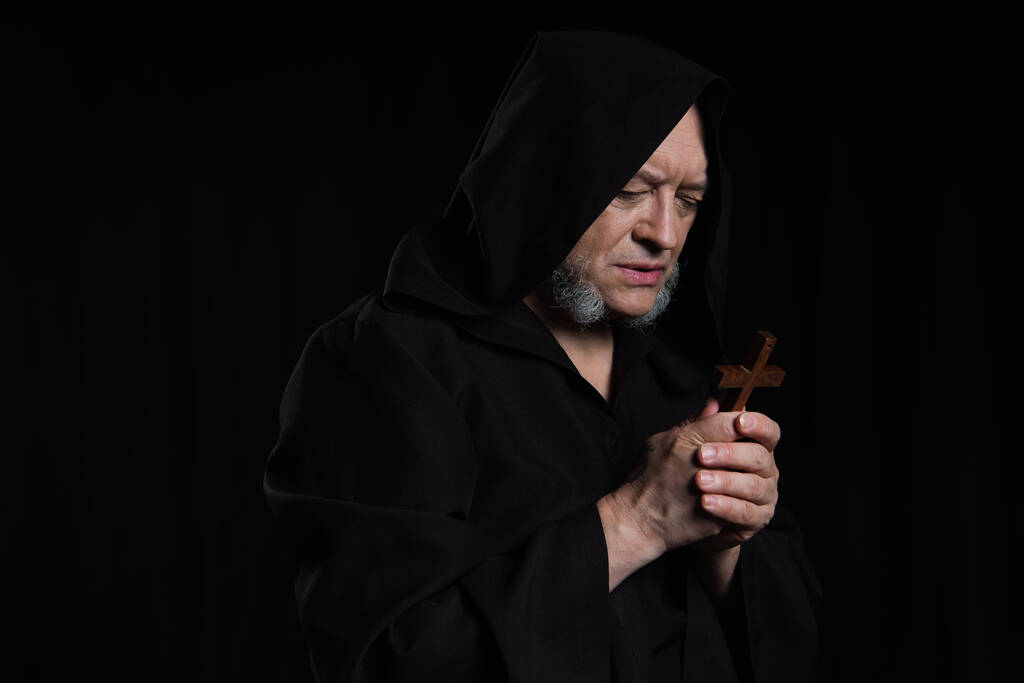monje mayor en la capucha mirando la cruz de madera mientras rezaba aislado en negro - Foto, Imagen