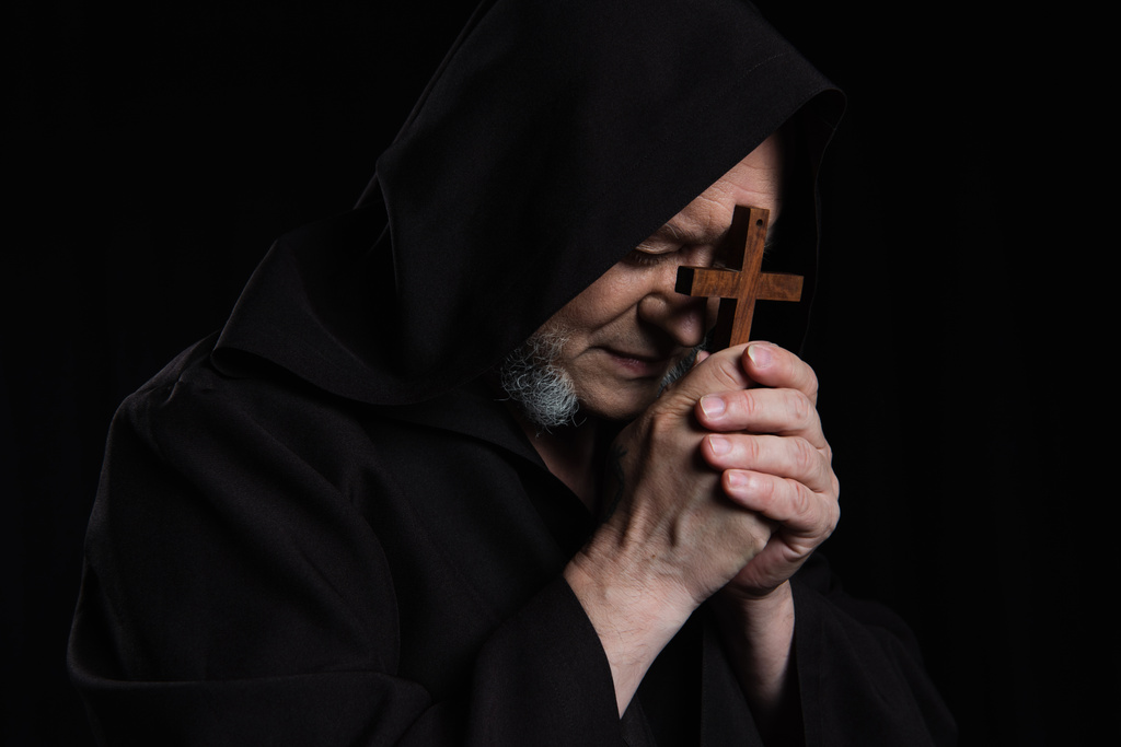 monje mayor en sotana encapuchada rezando con crucifijo cerca de la cara aislado en negro - Foto, imagen