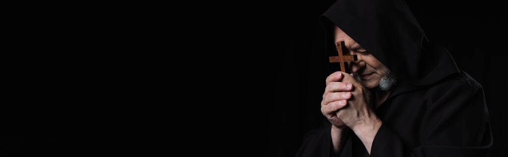 monaco anziano in cappuccio che tiene il crocifisso di legno vicino al volto e prega isolato sul nero, striscione - Foto, immagini