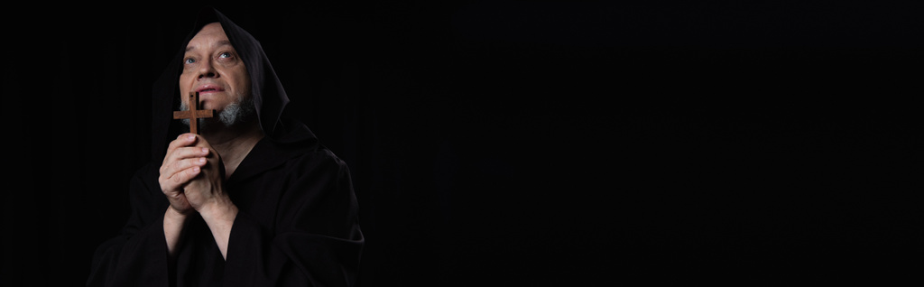 moine aîné en soutane priant avec une croix en bois et regardant isolé sur noir, bannière - Photo, image