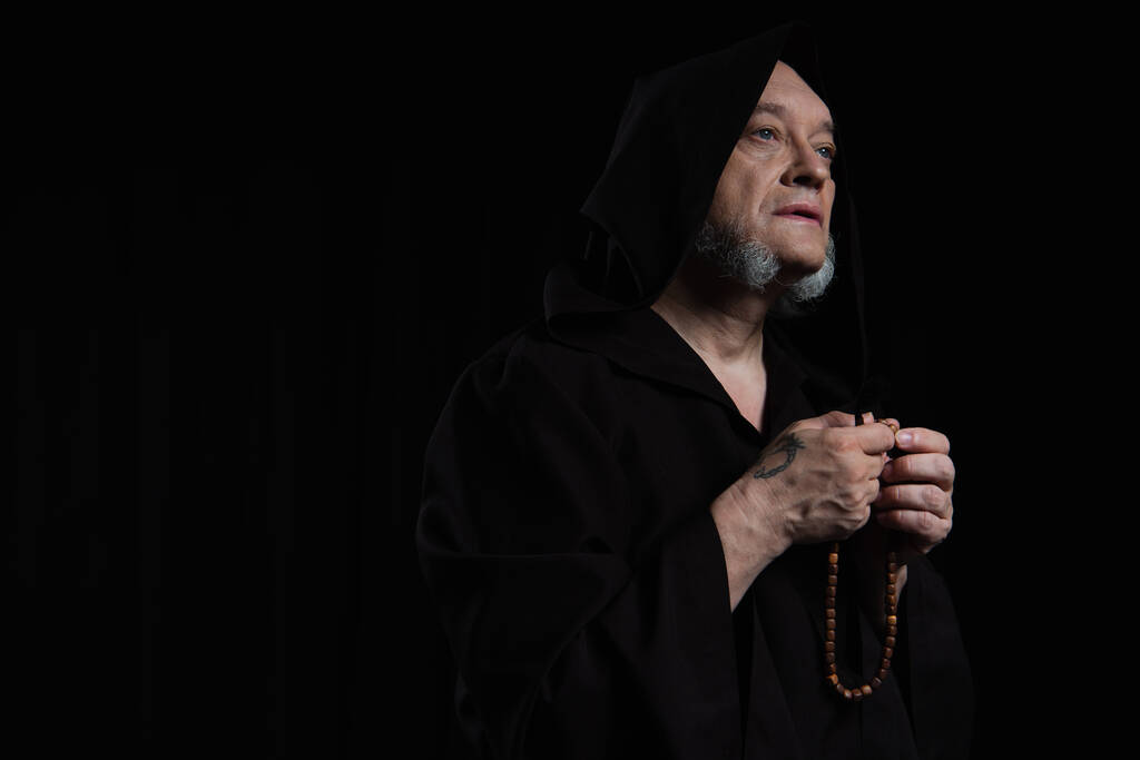 tattooed monk in hooded cassock praying with rosary isolated on black - Photo, Image