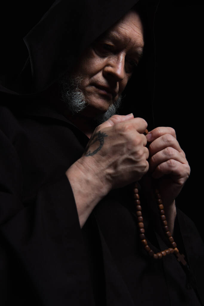 ανώτερος μοναχός με τατουάζ στο καπό κρατώντας κομπολόι, ενώ προσεύχεται απομονωμένη σε μαύρο - Φωτογραφία, εικόνα