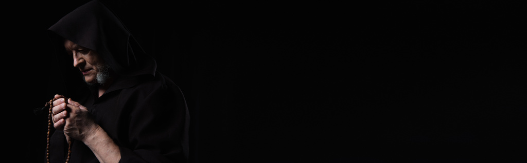 moine en soutane à capuchon foncé priant avec chapelet isolé sur noir, bannière - Photo, image