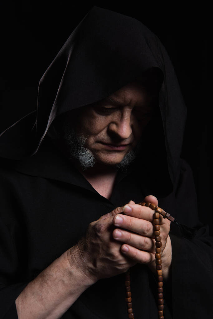 sacerdote medieval en bata encapuchada rezando con cuentas de rosario aisladas en negro - Foto, imagen