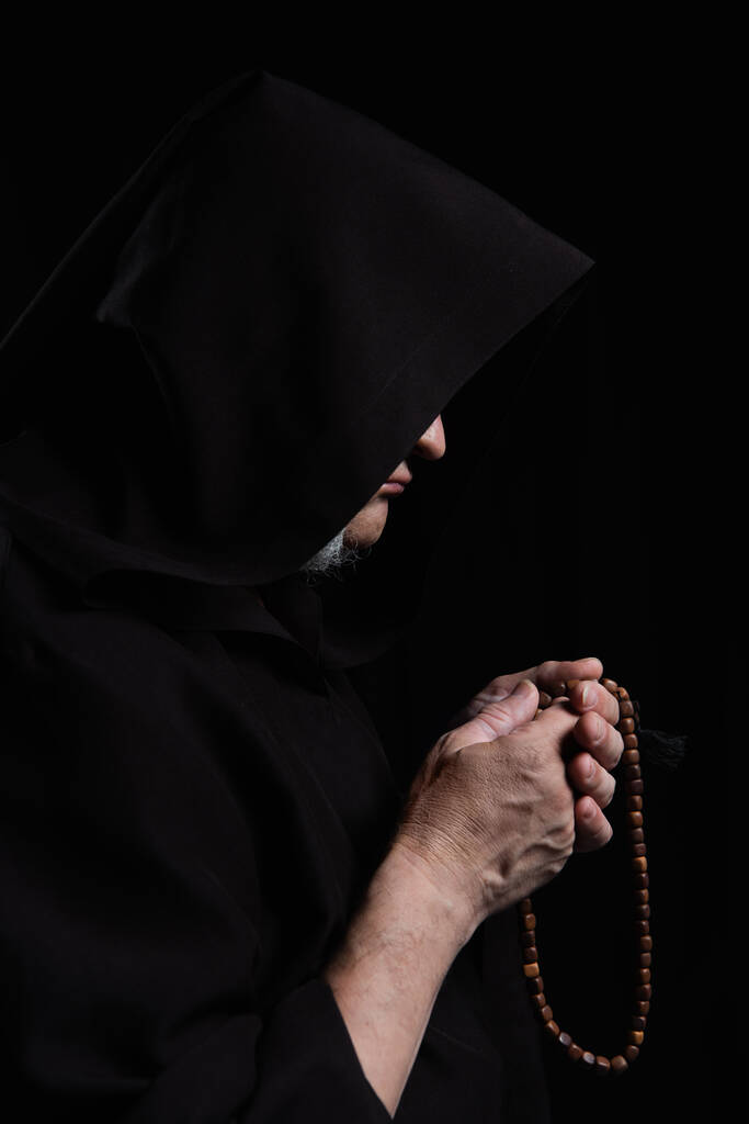 side view of mysterious monk with face obscured under hood praying with rosary isolated on black - Photo, Image