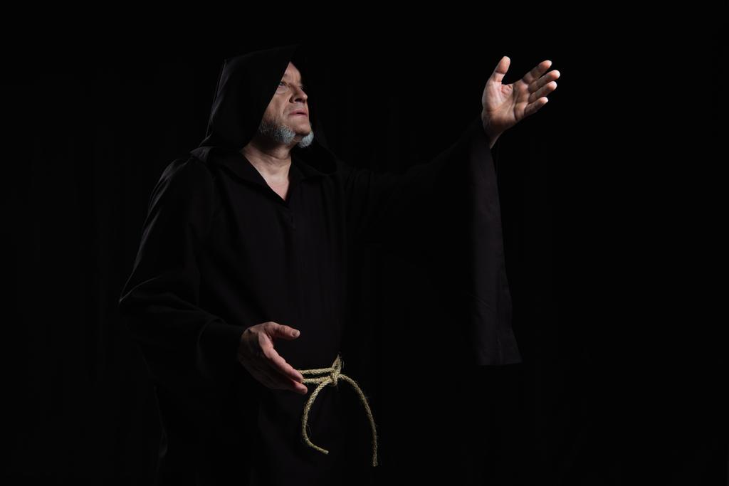 монах в рясе с капюшоном, указывая рукой, молясь изолированно на черном - Фото, изображение