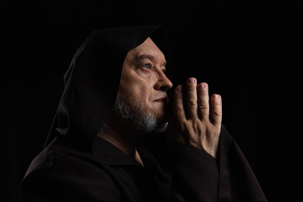 vista lateral del monje medieval con las manos de oración cerca de la cara aislada en negro - Foto, imagen