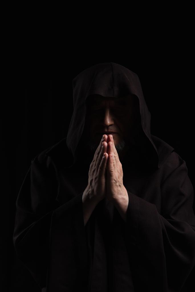 vooraanzicht van monnik in donker kapuchon soutane bidden geïsoleerd op zwart - Foto, afbeelding