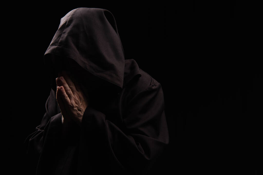 misterioso sacerdote con bata encapuchada rezando aislado en negro - Foto, imagen