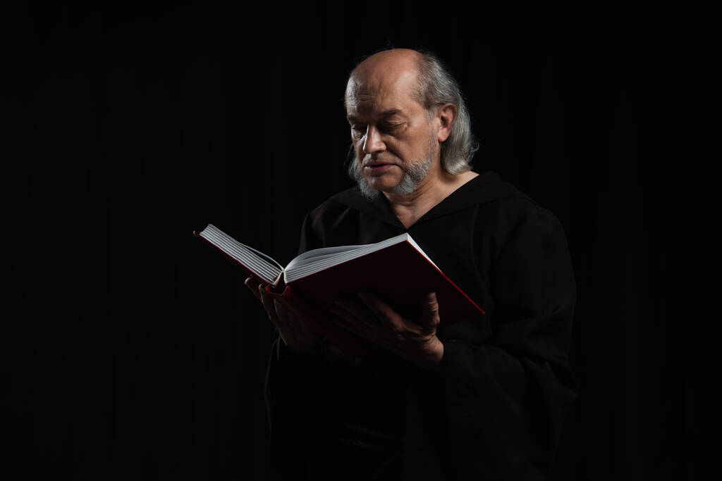 sacerdote medieval en túnica oscura leyendo la sagrada Biblia aislado en negro - Foto, Imagen