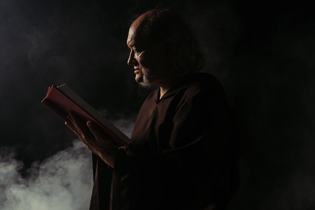 Cüppeli kıdemli rahibin siyah üzerine dumanlı kutsal İncil okuduğu yan manzara. - Fotoğraf, Görsel
