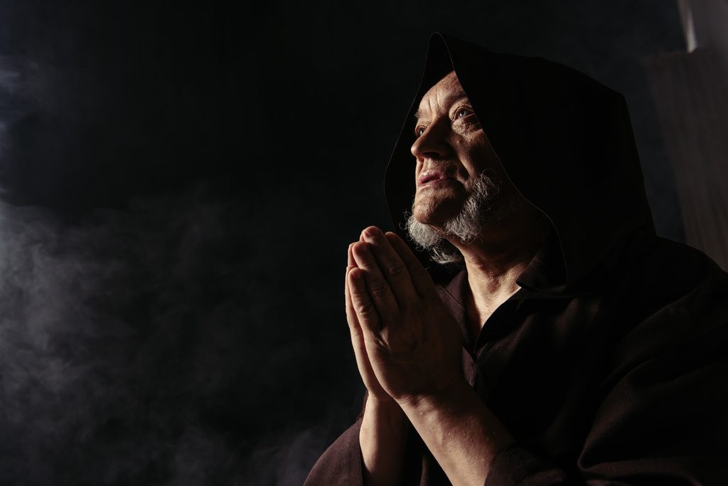 низкий угол зрения монаха в черном халате в капюшоне молящегося на темном фоне - Фото, изображение