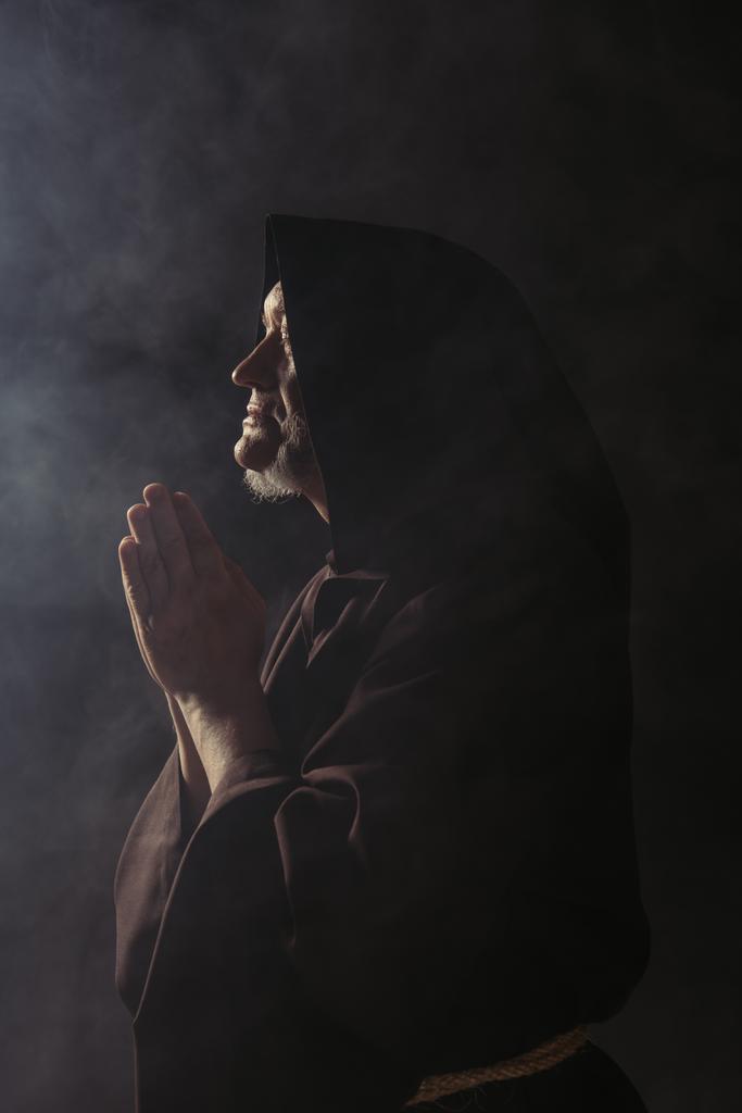 side view of medieval monk in hooded cassock praying on black with smoke - Photo, Image