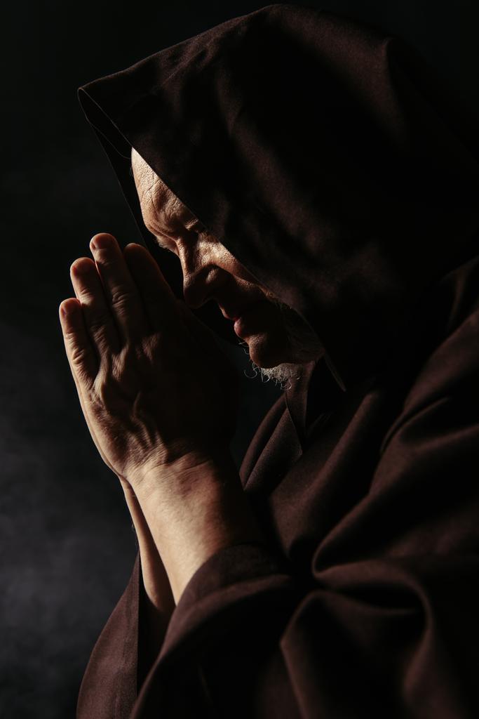 zijaanzicht van hoge priester met biddende handen en in zwarte kap op donkere achtergrond - Foto, afbeelding