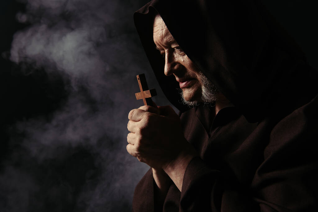 boční pohled na mnicha v černé kápi sutaně modlení se svatým křížem na tmavém pozadí s kouřem - Fotografie, Obrázek