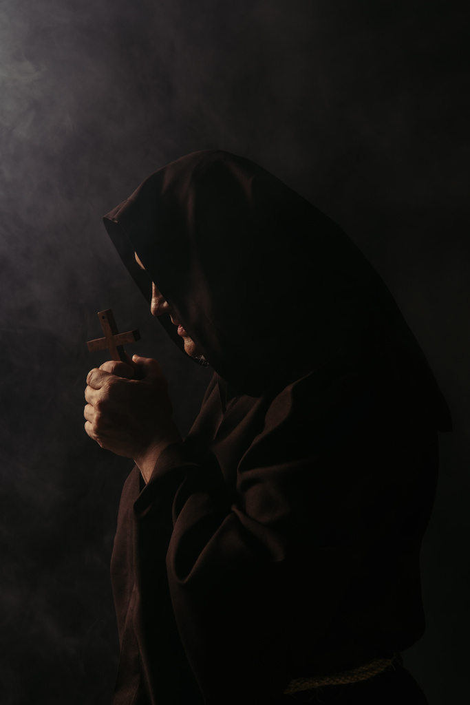フード付きのローブを着た謎の中世の僧が黒を背景に十字架で祈る - 写真・画像