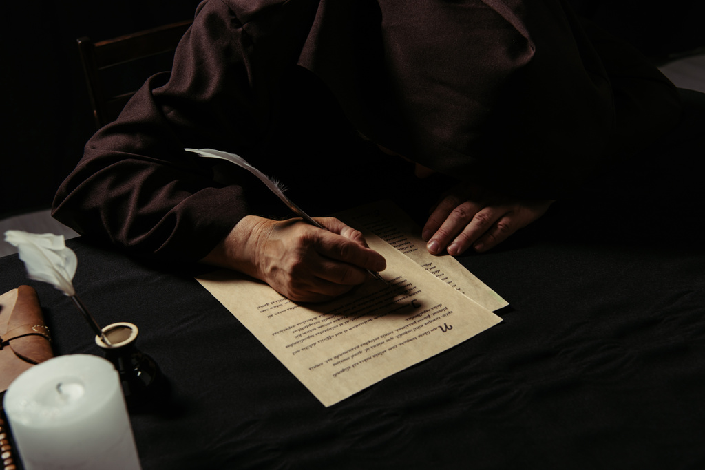 средневековый монах со склоненной головой пишет рукопись на пергаменте, изолированном на черном - Фото, изображение