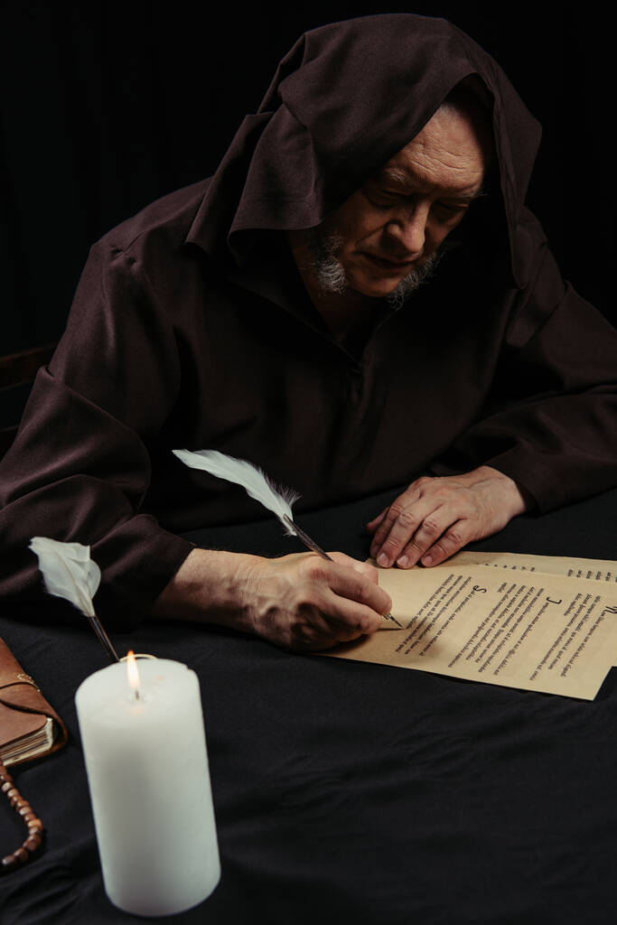 monaco in accappatoio incappucciato scritto su pergamena vicino candela accesa di notte isolato su nero - Foto, immagini