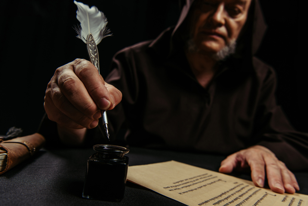 blurred monk holding feather pen near inkpot and ancient manuscript isolated on black - Photo, Image