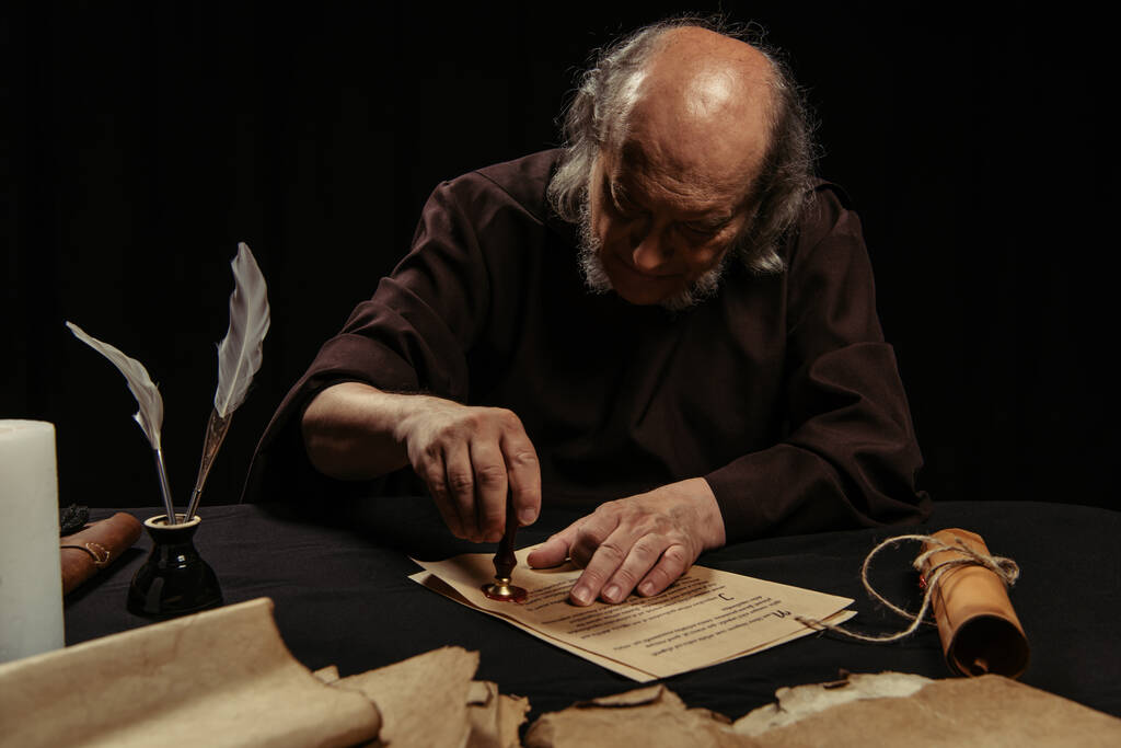 старший монах печатает рукопись с восковой печатью возле пергаментов, изолированных на черном - Фото, изображение