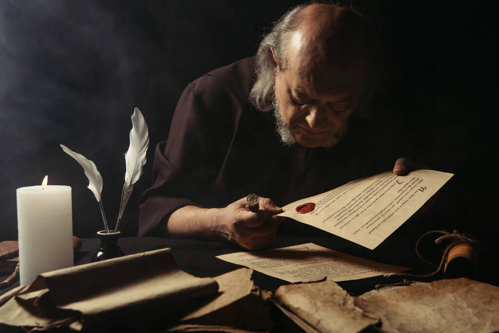 senior monk looking at chronicle stamped with wax seal near parchments and burning candle on dark background - Photo, Image