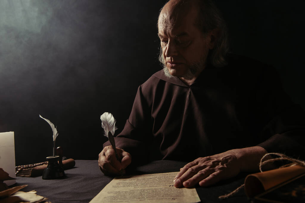 medieval priest in black robe writing manuscript on dark background - Photo, Image