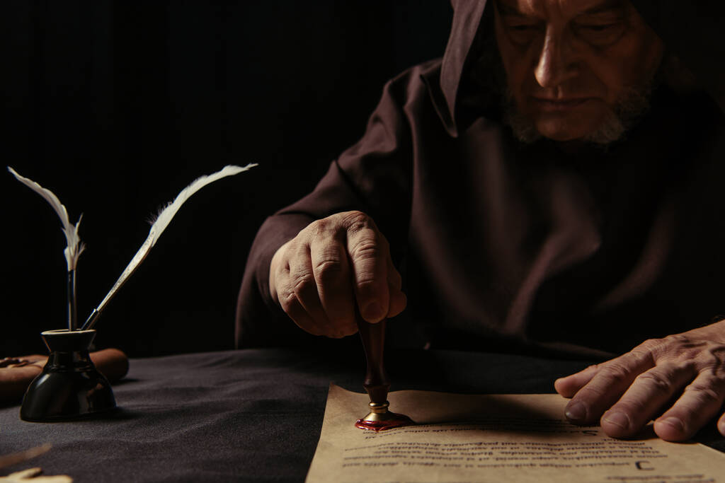 Средневековый монах в темной одежде с вышивкой на капюшоне на пергаменте с восковой печатью, изолированной на черном - Фото, изображение