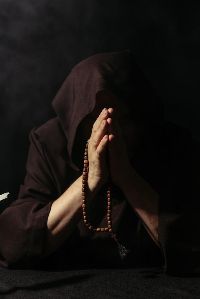 anonieme monnik in capuchon badjas bidden met rozenkrans kralen geïsoleerd op zwart - Foto, afbeelding