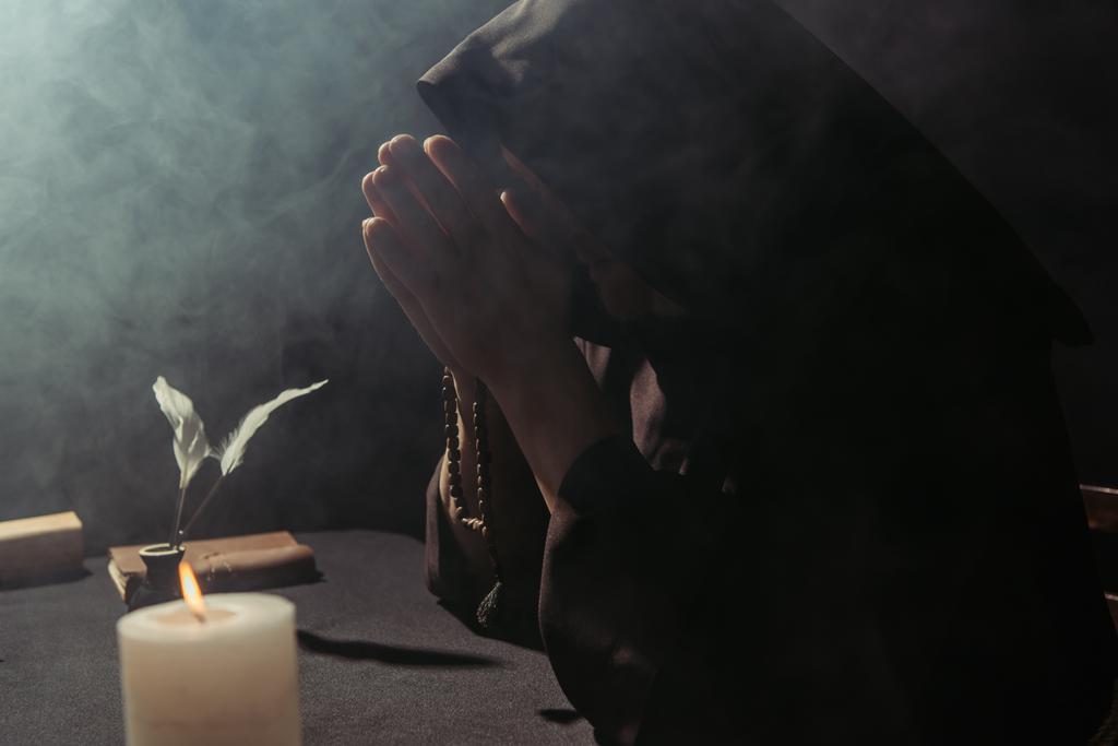 中世の僧でフード付きのキャソックで祈ります煙と黒の背景に燃えるろうそくの近くにバラ色のビーズ - 写真・画像
