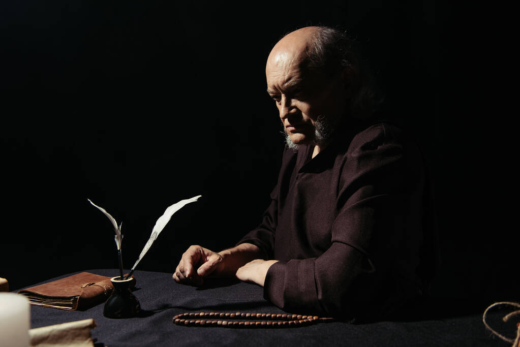 monge pensativo sentado perto de tinteiro e rosário à noite isolado em preto - Foto, Imagem