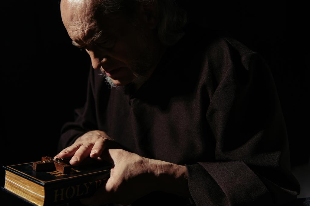 monk in dark robe holding bible and crucifix isolated on black - Photo, Image