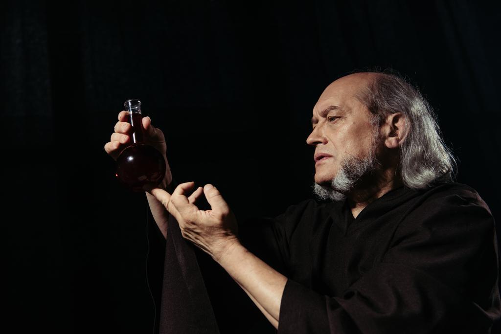 vousatý alchymista v plášti drží baňku s lektvarem izolované na černé - Fotografie, Obrázek