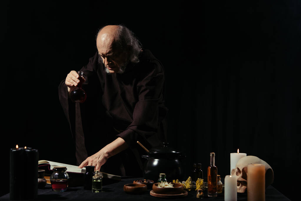 średniowieczny uzdrowiciel trzymając piersiówkę i czytając magiczną książkę kucharską w pobliżu ziół odizolowanych na czarno - Zdjęcie, obraz
