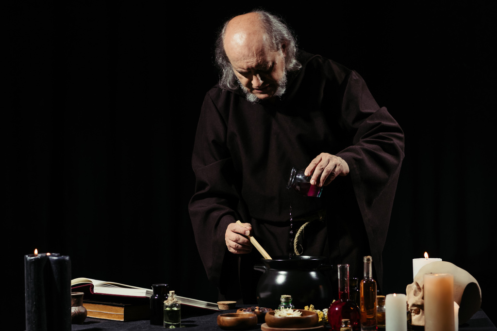 alquimista medieval preparando poción mágica mientras añade ingrediente en maceta aislada en negro - Foto, imagen