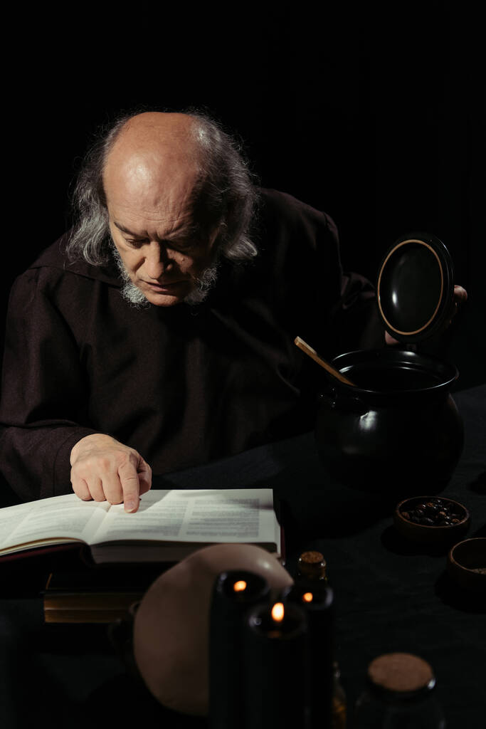 senior alchemist reading magic cookbook near pot isolated on black - Photo, Image