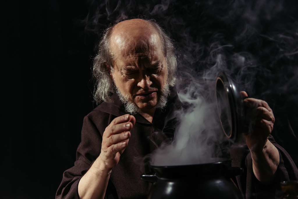 Senior Alchemist runzelt die Stirn in der Nähe dampfenden Topf isoliert auf schwarz - Foto, Bild