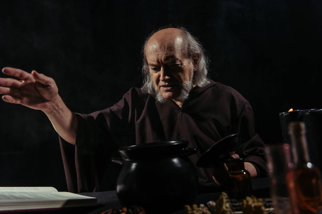 таємничий алхімік з простягнутою рукою біля горщика і чарівною кулінарною книгою ізольовані на чорному
 - Фото, зображення