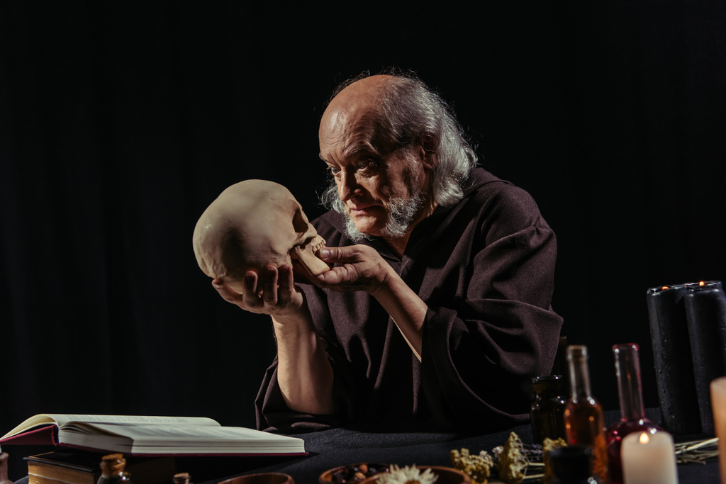 魔法の料理本の近くで人間の頭蓋骨を見る神秘的な錬金術師と黒で隔離された乾燥ハーブ - 写真・画像
